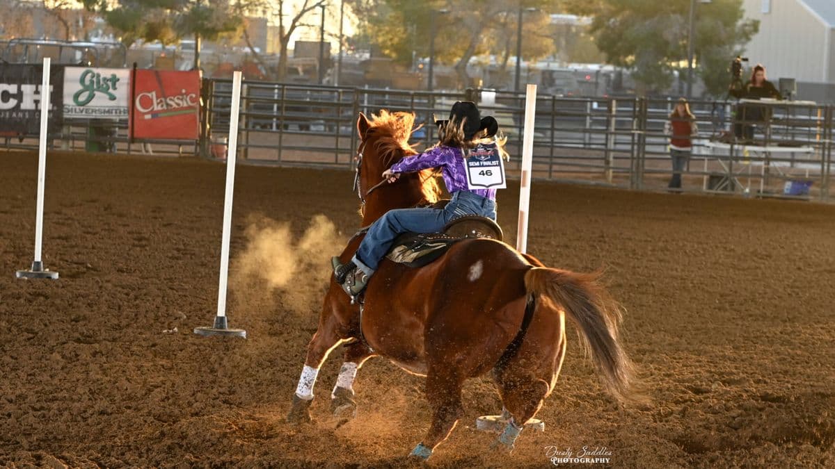 Young rider on a brown horse navigating a barrel racing course at a rodeo arena, kicking up dust as they approach a white pole marker