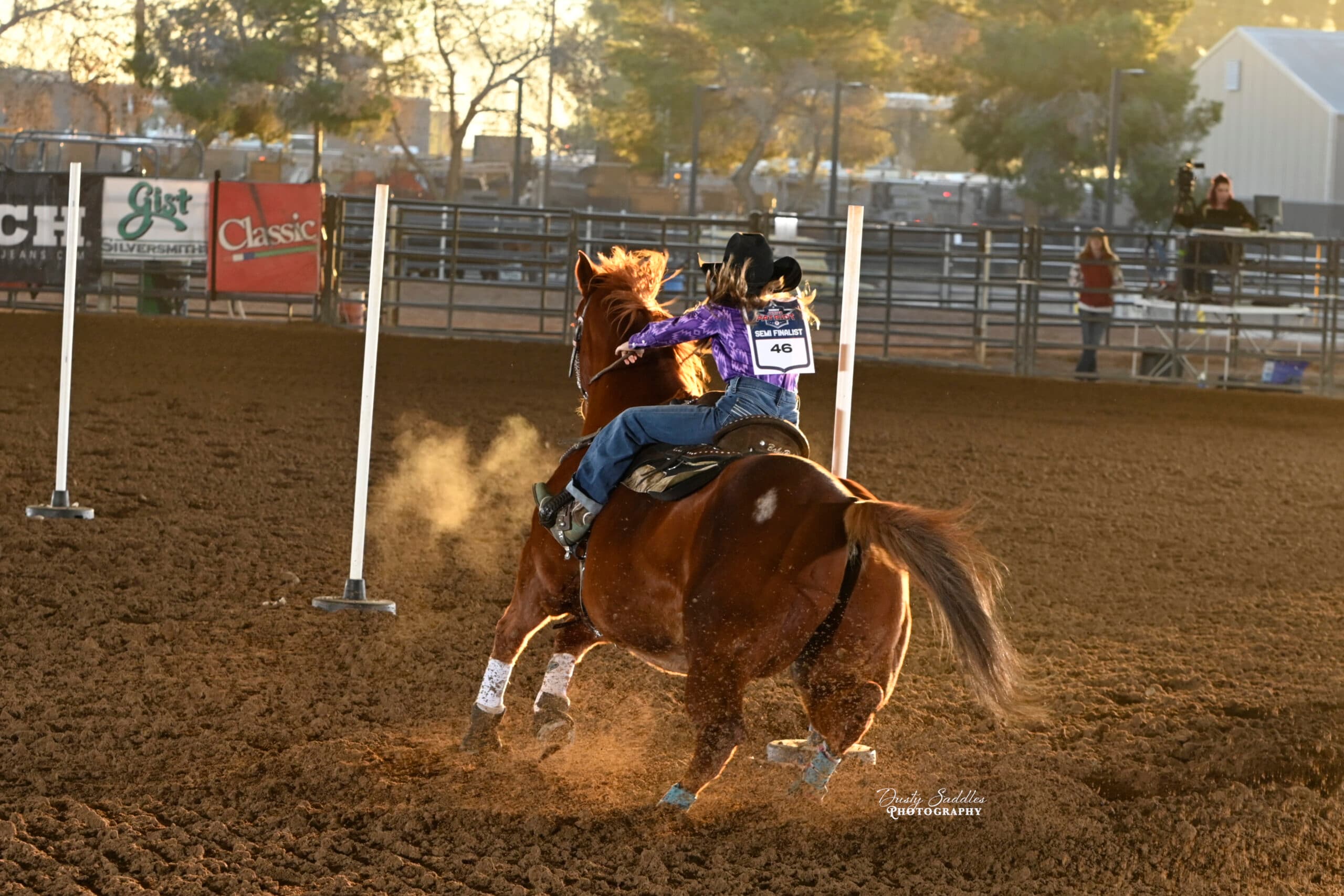 Young rider on a brown horse navigating a barrel racing course at a rodeo arena, kicking up dust as they approach a white pole marker