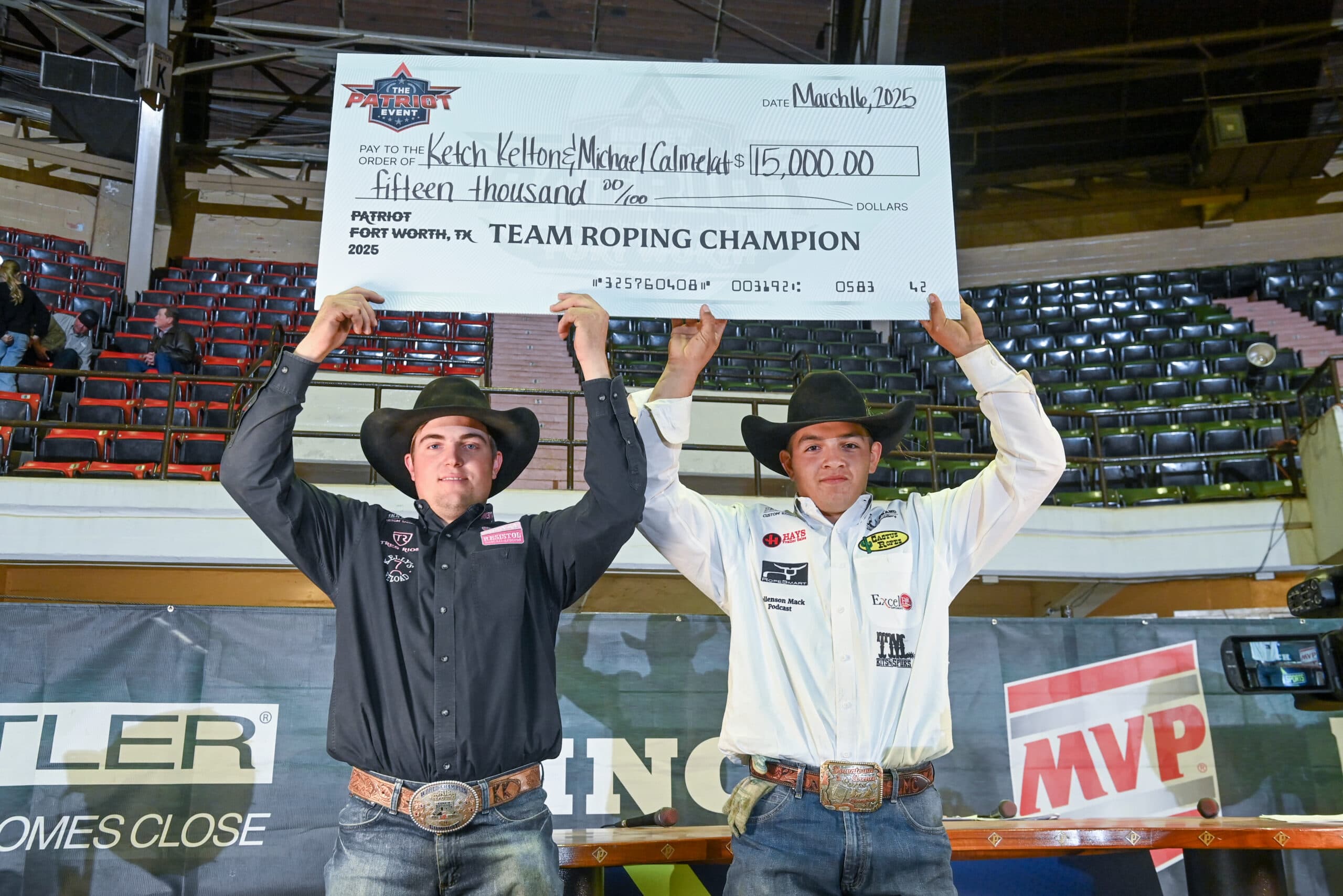 Two team roping champions hold a large ceremonial check for $15,000 at the 2025 Patriot Fort Worth event in an indoor arena