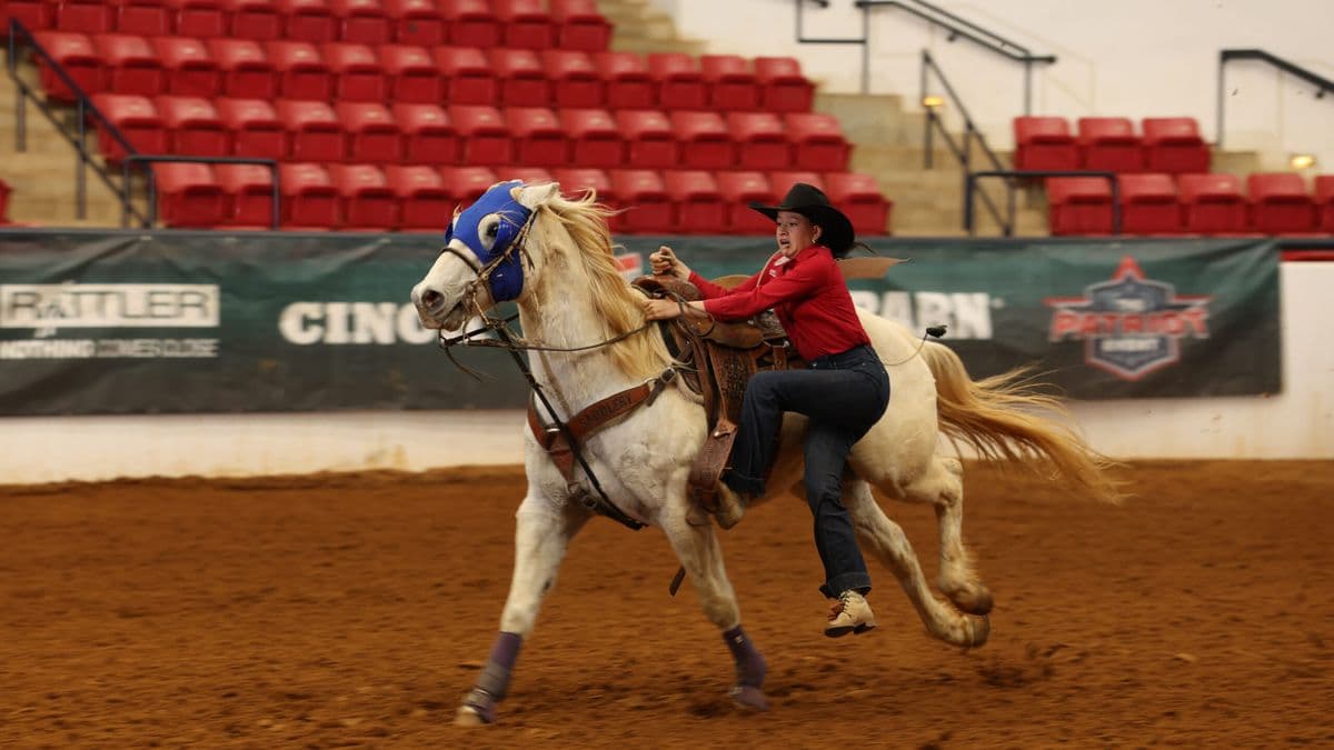 Barrel racer in red shirt and black cowboy hat rides palomino horse across dirt arena during rodeo competition