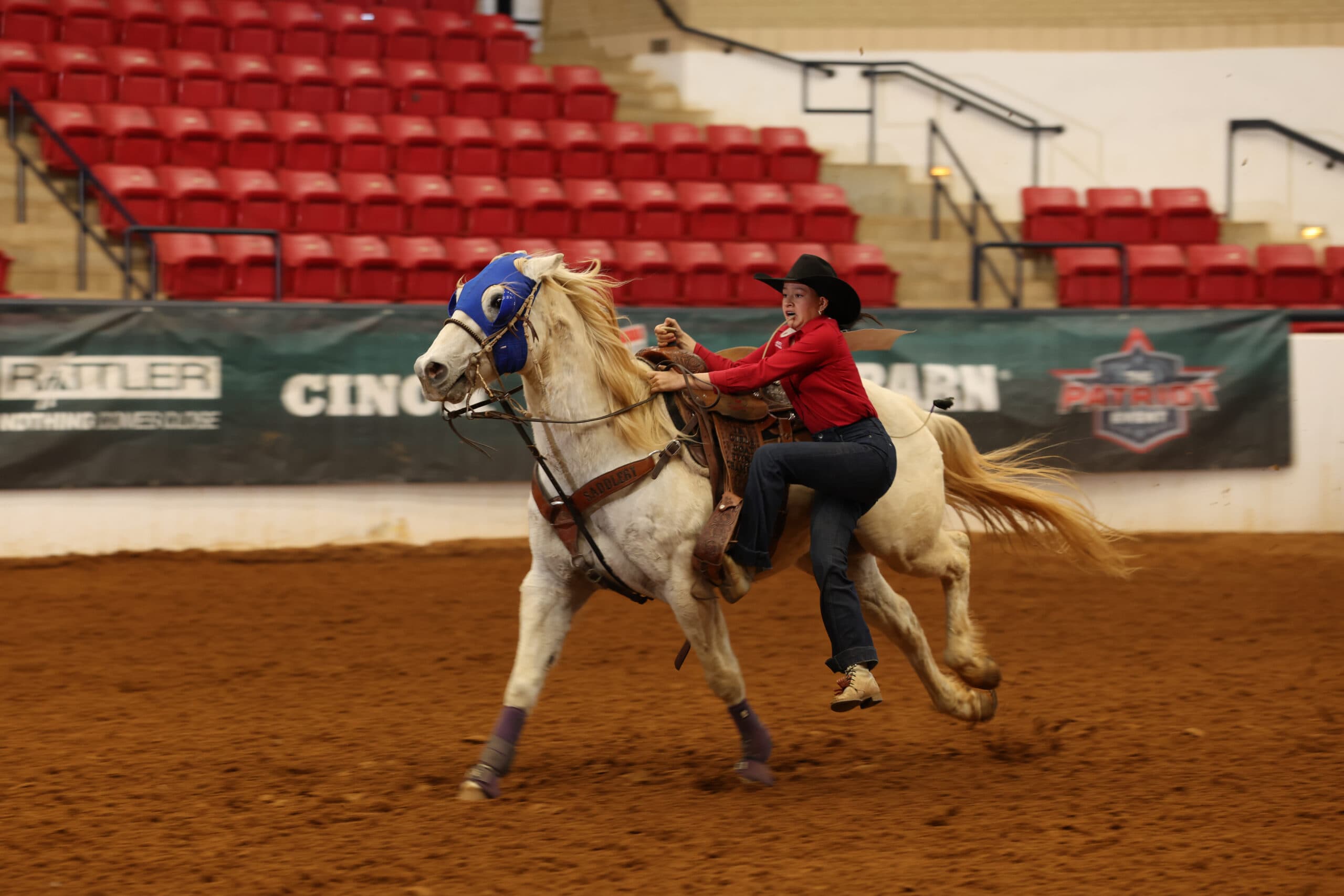 Barrel racer in red shirt and black cowboy hat rides palomino horse across dirt arena during rodeo competition