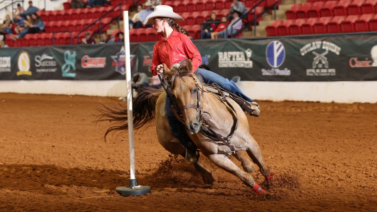 Barrel racer in red shirt and white cowboy hat rounds a pole on brown horse at indoor rodeo arena with spectators in stands