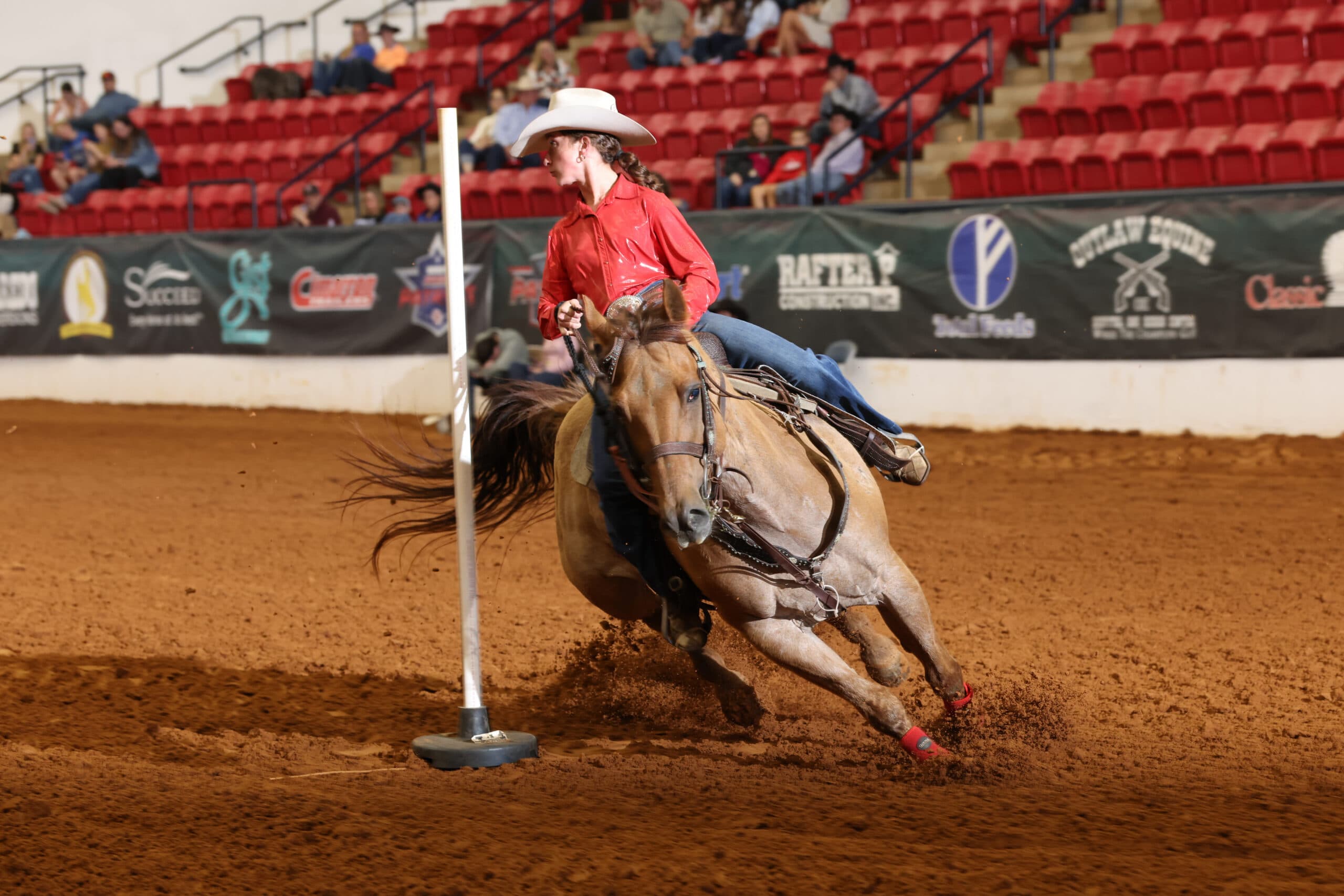 Barrel racer in red shirt and white cowboy hat rounds a pole on brown horse at indoor rodeo arena with spectators in stands