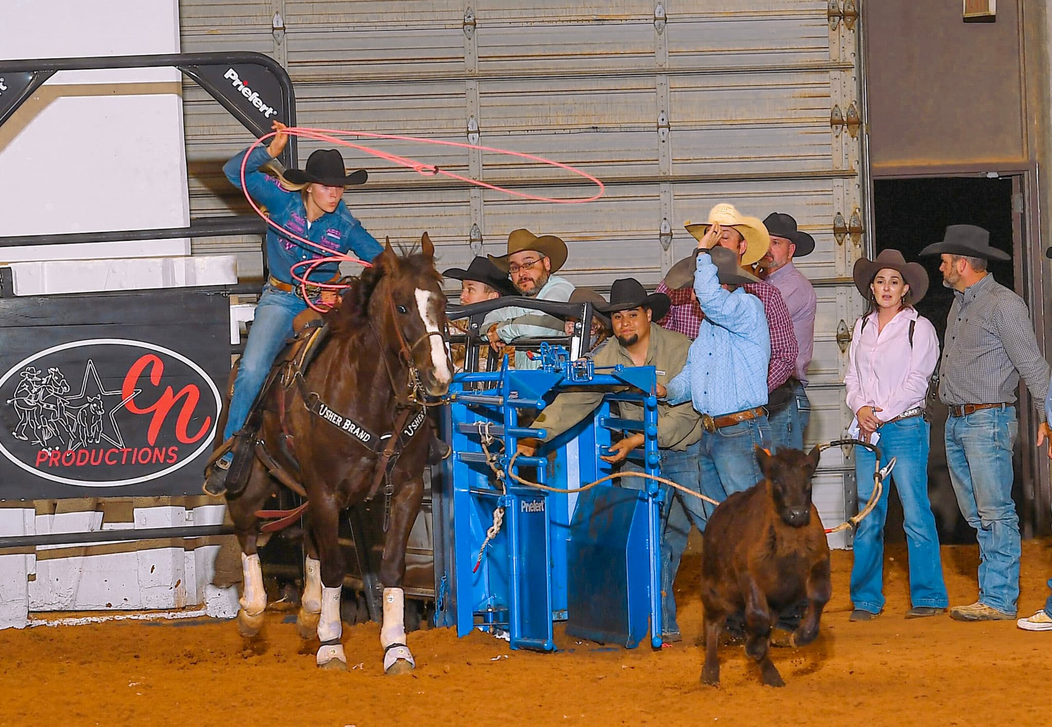 Roper on horseback swings lasso while team waits by blue chute at indoor rodeo event for Isher Brand Productions