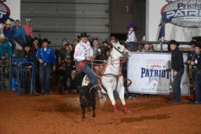 Cowboy on white horse roping steer during rodeo event at indoor arena with spectators and Patriot banner visible