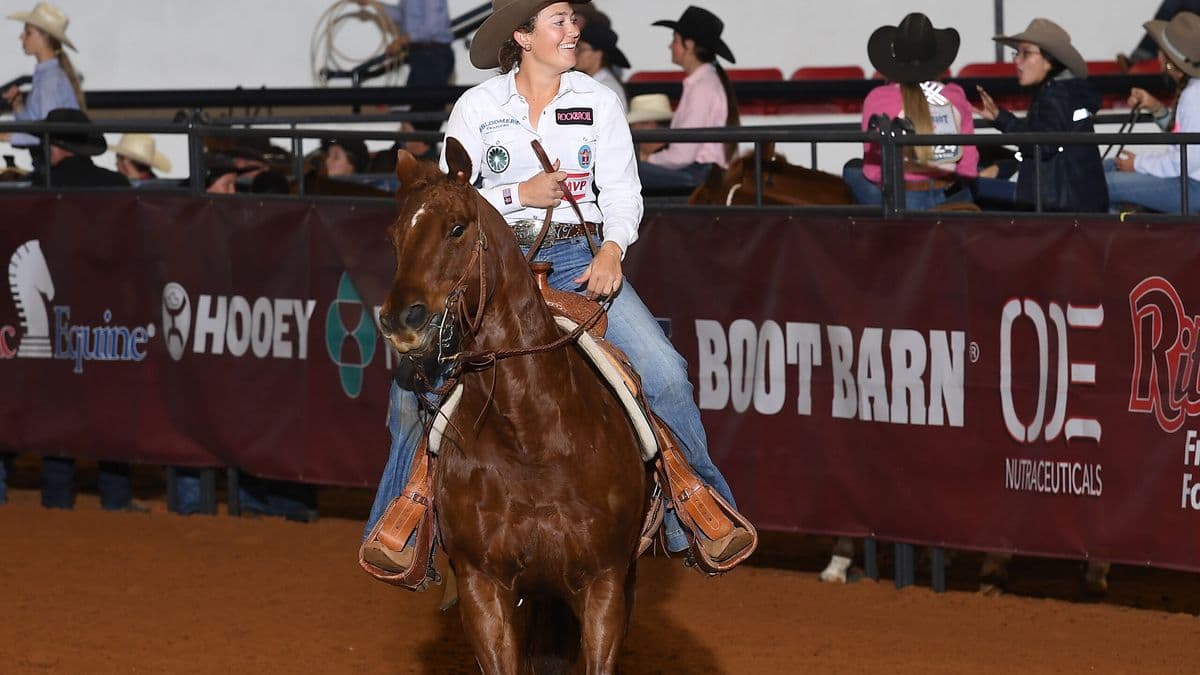 Rodeo competitor on brown horse rides past sponsor barrier at indoor arena event, wearing white shirt and cowboy hat
