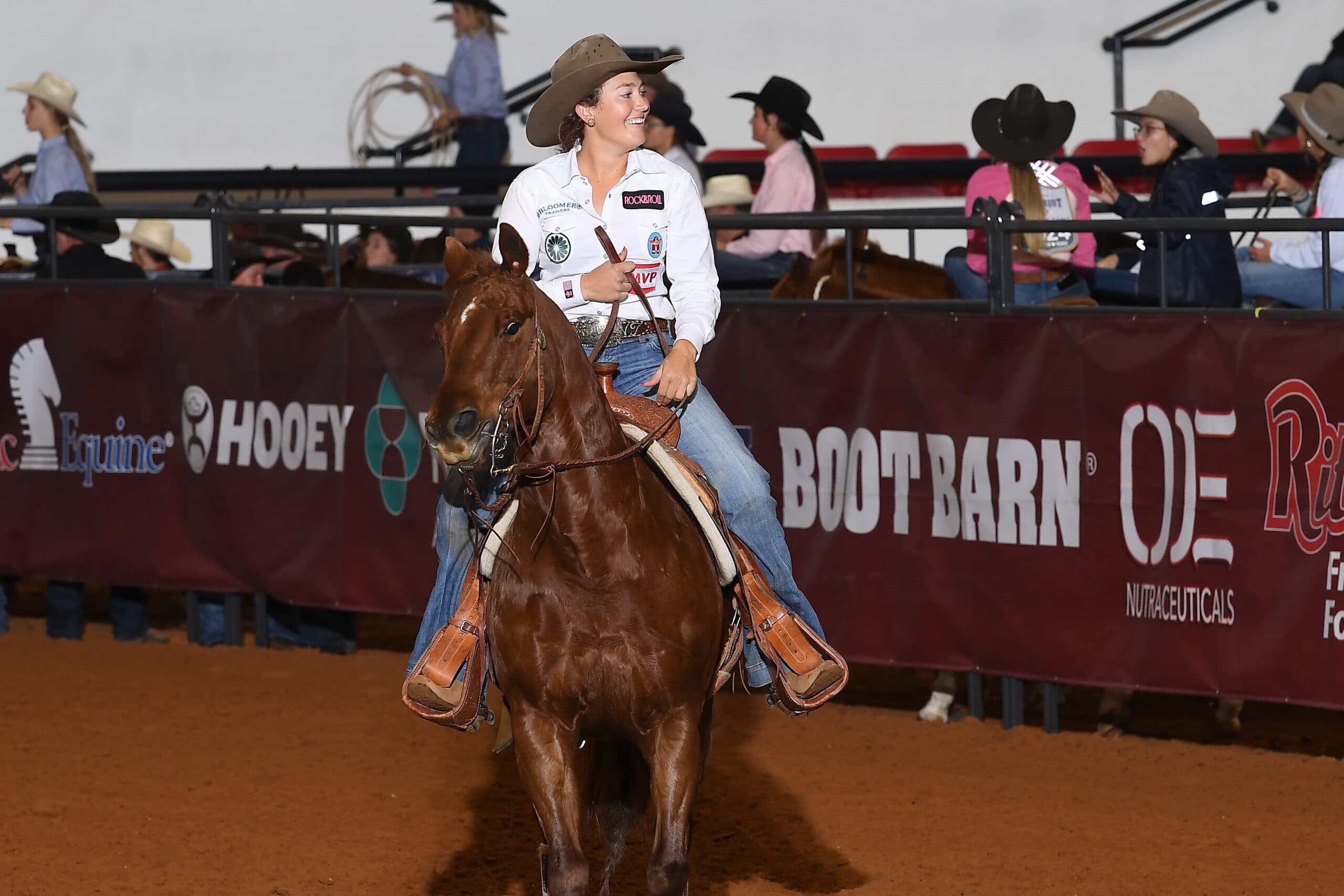 Rodeo competitor on brown horse rides past sponsor barrier at indoor arena event, wearing white shirt and cowboy hat