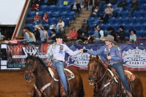 Two mounted cowboys on horseback ride across a dirt arena at a rodeo with blue stadium seating in the background