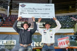 Two rodeo competitors hold a large ceremonial check above their heads at an indoor arena, celebrating a victory