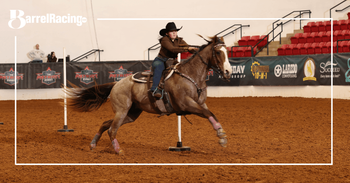 Barrel racer on brown horse turning around barrel during competition in indoor arena with red stadium seating