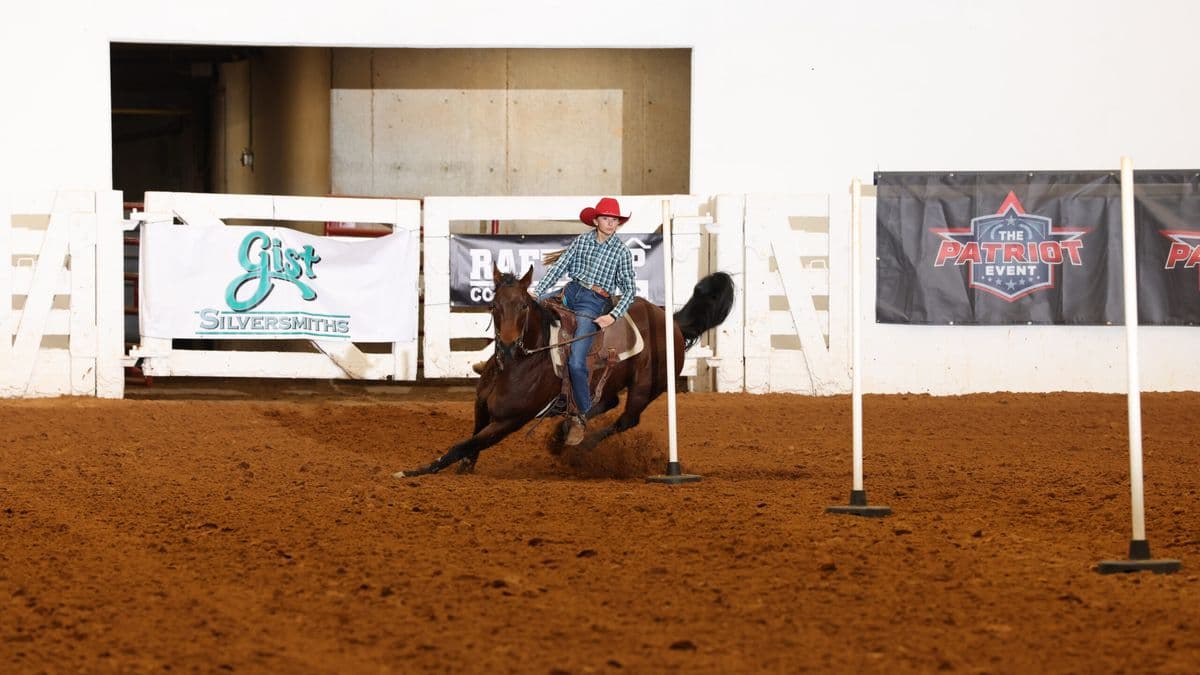 Barrel racer on brown horse rounds a barrel during competition at The Patriot Event, with Gist Silversmiths signage visible in background