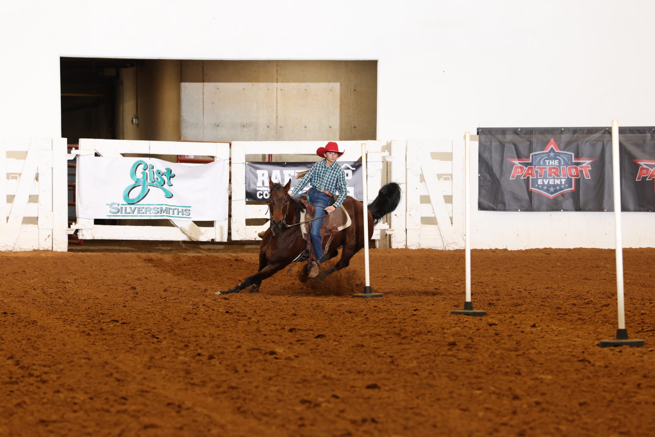 Barrel racer on brown horse rounds a barrel during competition at The Patriot Event, with Gist Silversmiths signage visible in background