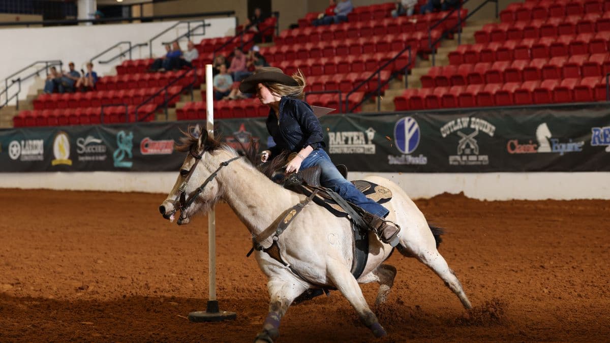 Barrel racer on white horse rounds a barrel during indoor rodeo competition with spectators in red seats behind