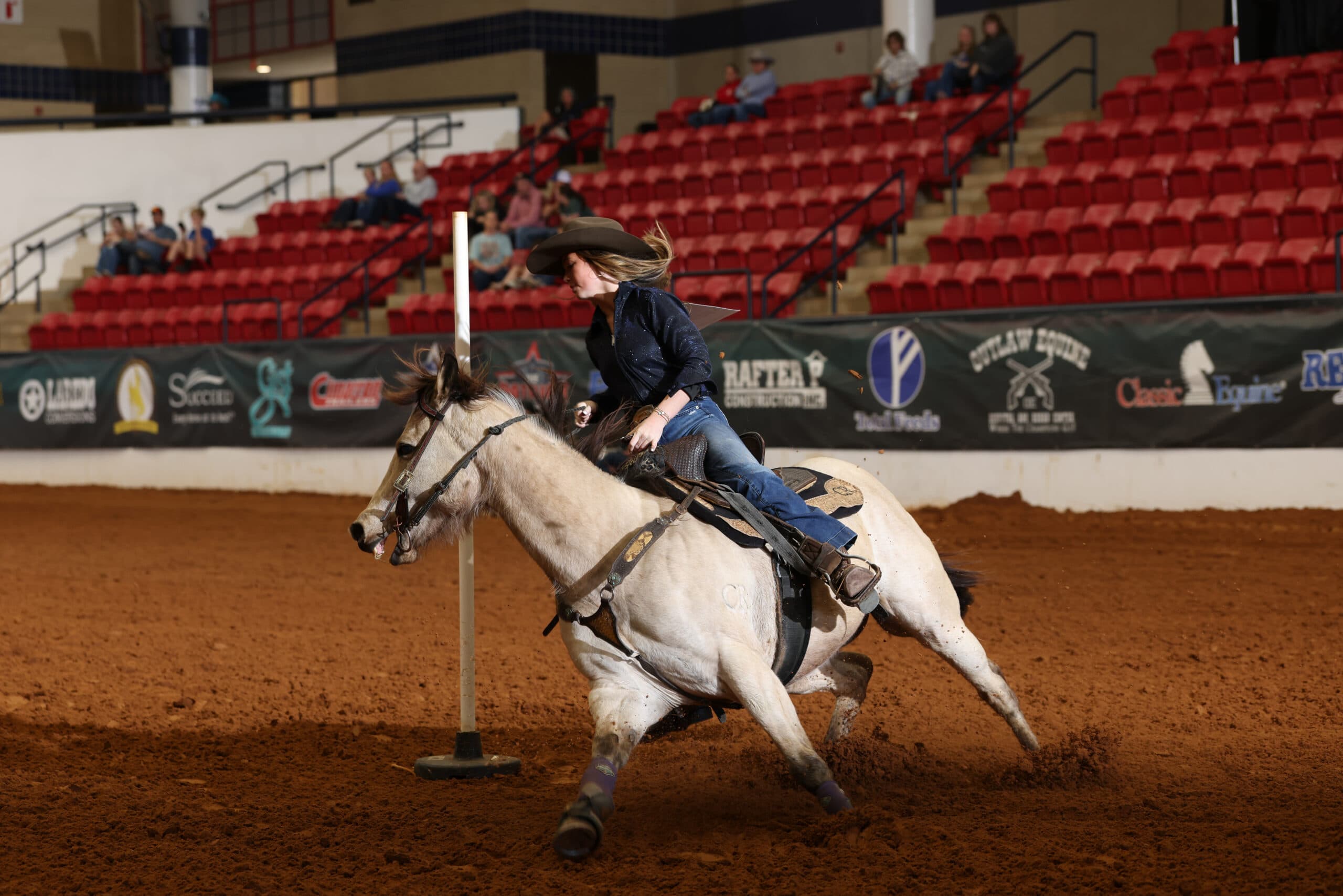 Barrel racer on white horse rounds a barrel during indoor rodeo competition with spectators in red seats behind