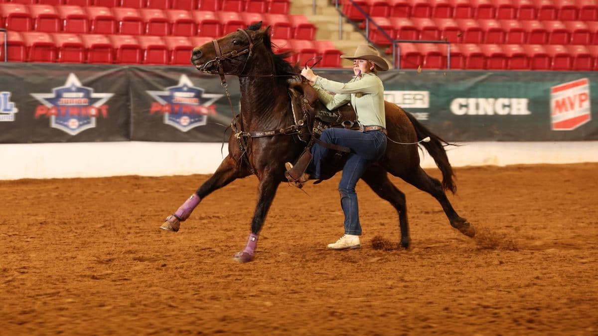 Cowgirl in tan shirt and cowboy hat rides dark horse with purple leg wraps in indoor arena during rodeo event
