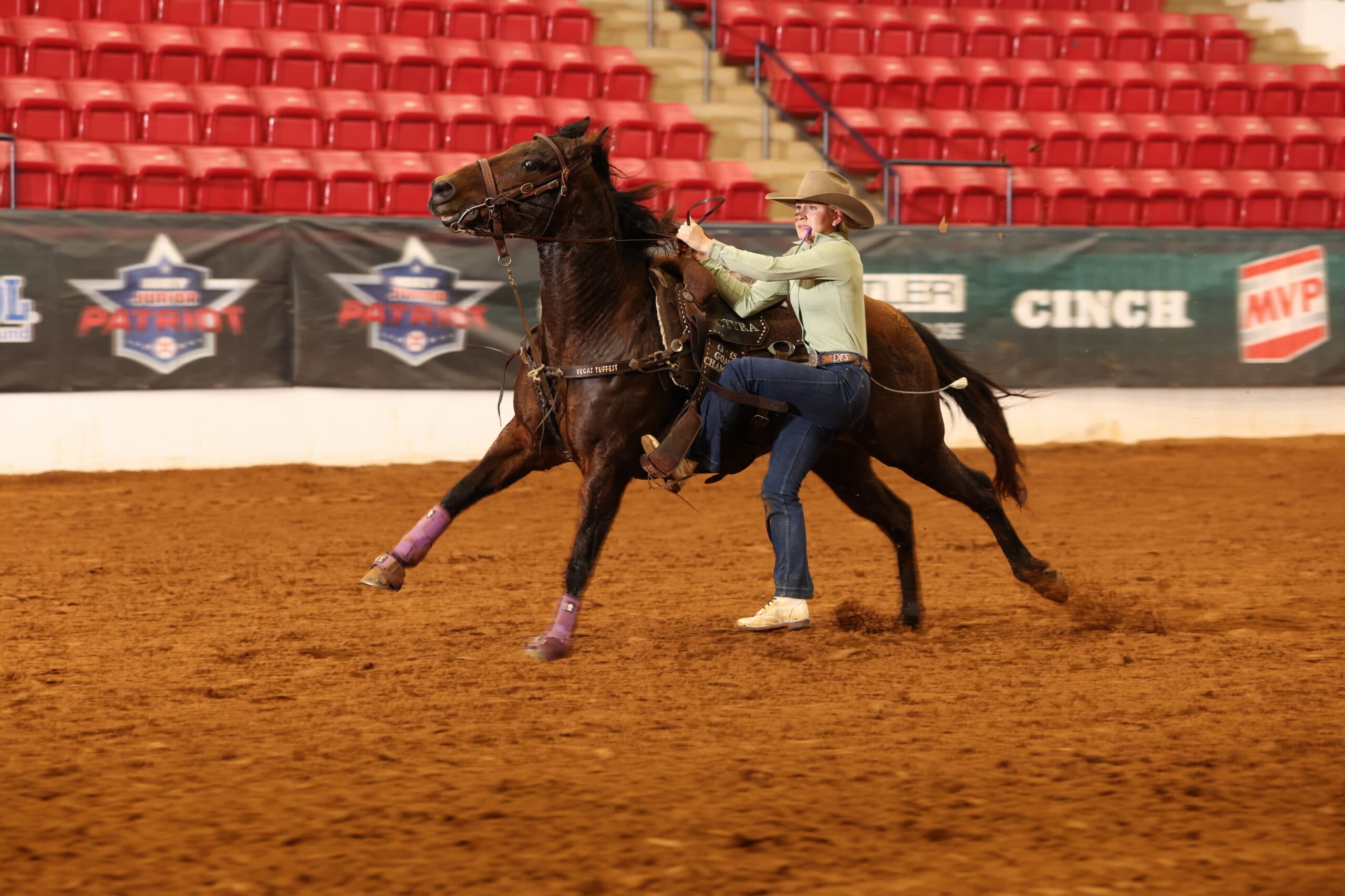 Cowgirl in tan shirt and cowboy hat rides dark horse with purple leg wraps in indoor arena during rodeo event